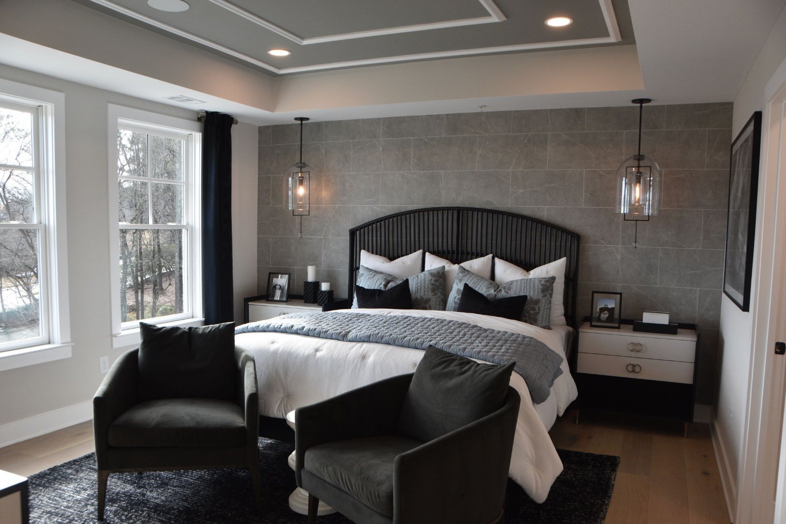 A staged primary bedroom ready for the next arrival: a slatted headboard against a stone feature wall, layered pillows, a quilted throw folded across the foot of the bed, glass pendant lights hanging either side, two armchairs at the foot on a dark rug, and daylight through tall windows at frame left